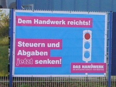 An einem Zaun hängendes Plakat mit Ampel in den Farben Rot-Blau-Blau und folgendem Text: „Dem Handwerk reichts! Steuern und Abgaben jetzt senken! DAS HANDWERK, DIE WIRTSCHAFTSMACHT VON NEBENAN“