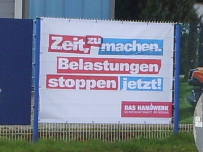 An einem Zaun hängendes Plakat mit folgendem Text: „Zeit, zu machen. Belastungen stoppen jetzt! DAS HANDWERK“