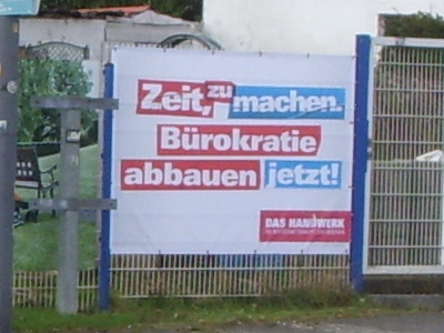 An einem Zaun hängendes Plakat mit folgendem Text: „Zeit, zu machen. Bürokratie abbauen jetzt! DAS HANDWERK“