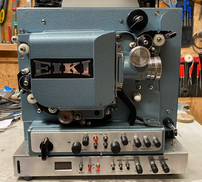 Historisch
	aussehender Schmalfilm Projektor mit Markenlogo EIKI, der auf ein
	modernes Steuerpult aufgeschraubt ist, eingeklappt in einer Werkstatt
	stehend.